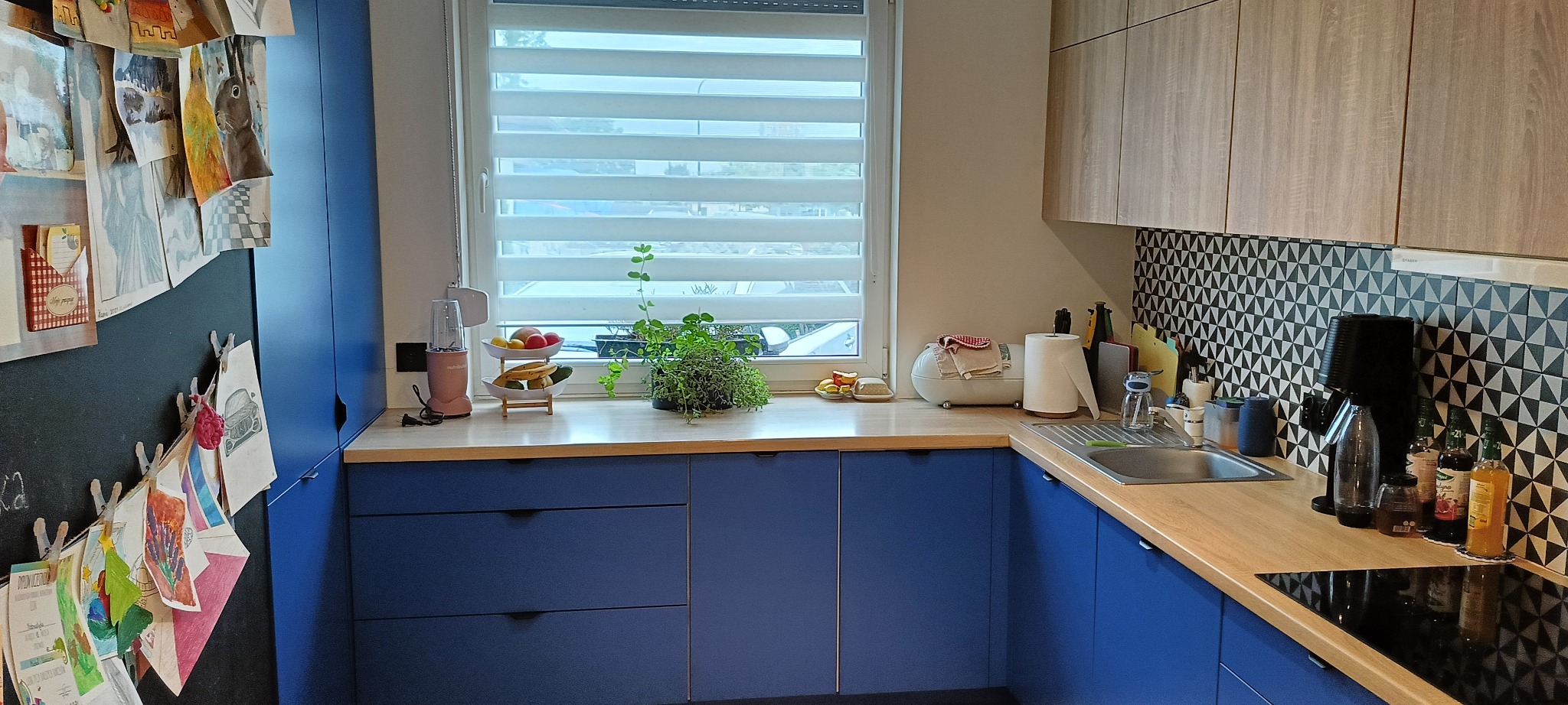 Niebieska kuchnia z drewnianym blatem, wzorzystą ścianą i oknem z żaluzjami. Na blacie akcesoria kuchenne i dekoracje. Na ścianie rysunki dziecięce.