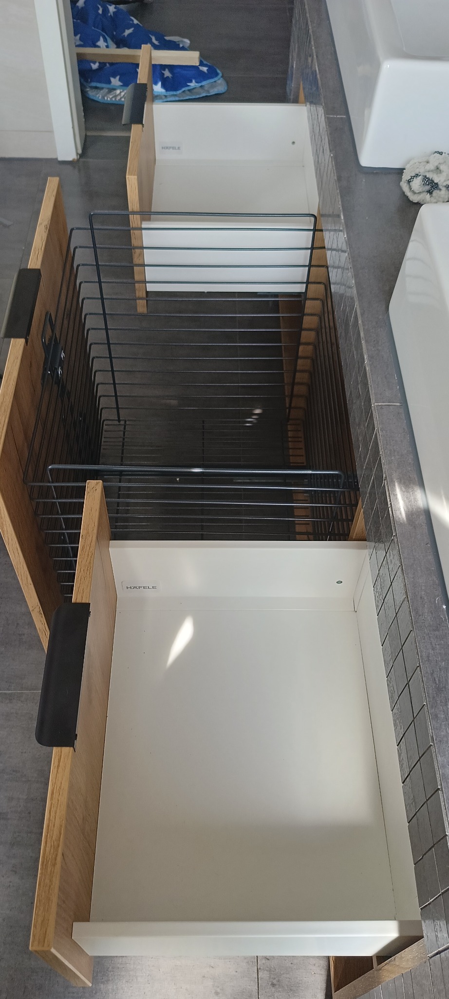 Otwarta szafka łazienkowa z drewnianymi frontami i metalowym koszem na akcesoria, widok z góry, z widocznym logo Hafele na wewnętrznej stronie szuflady.