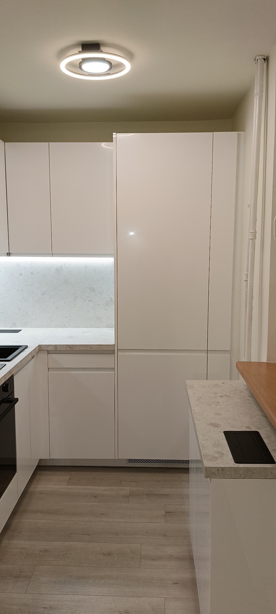 Biała, minimalistyczna kuchnia na wymiar z oświetleniem LED pod szafkami i okrągłą lampą sufitową. Blat imitujący kamień. Widoczna wentylacja pod szafkami.