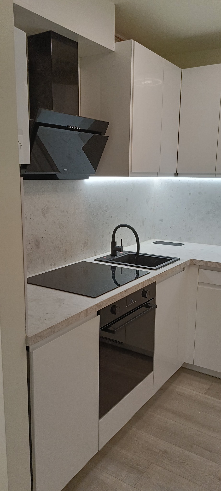 Biała, nowoczesna kuchnia z czarnym zlewem, płytą indukcyjną i piekarnikiem. Uwagę przyciąga minimalistyczny okap i subtelne oświetlenie LED pod szafkami.