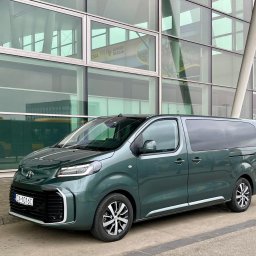 PIOTRANSPORT - Piotr Pacha - Zielony van Toyota Proace Verso z przyciemnianymi szybami, zaparkowany przed nowoczesnym budynkiem z przeszkloną fasadą. Tablica rejestracyjna widoczna.