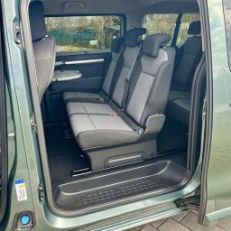 PIOTRANSPORT - Piotr Pacha - Wnętrze zielonego busa z otwartymi drzwiami suwanymi, widoczne trzy rzędy siedzeń z szarą tapicerką i stolik. Podłoga wyłożona czarną wykładziną.