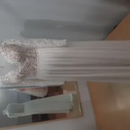 Elegancka suknia ślubna z długimi rękawami i koronkową górą, widoczna na manekinie z odbiciem w lustrze, na tle ściany i blatu.