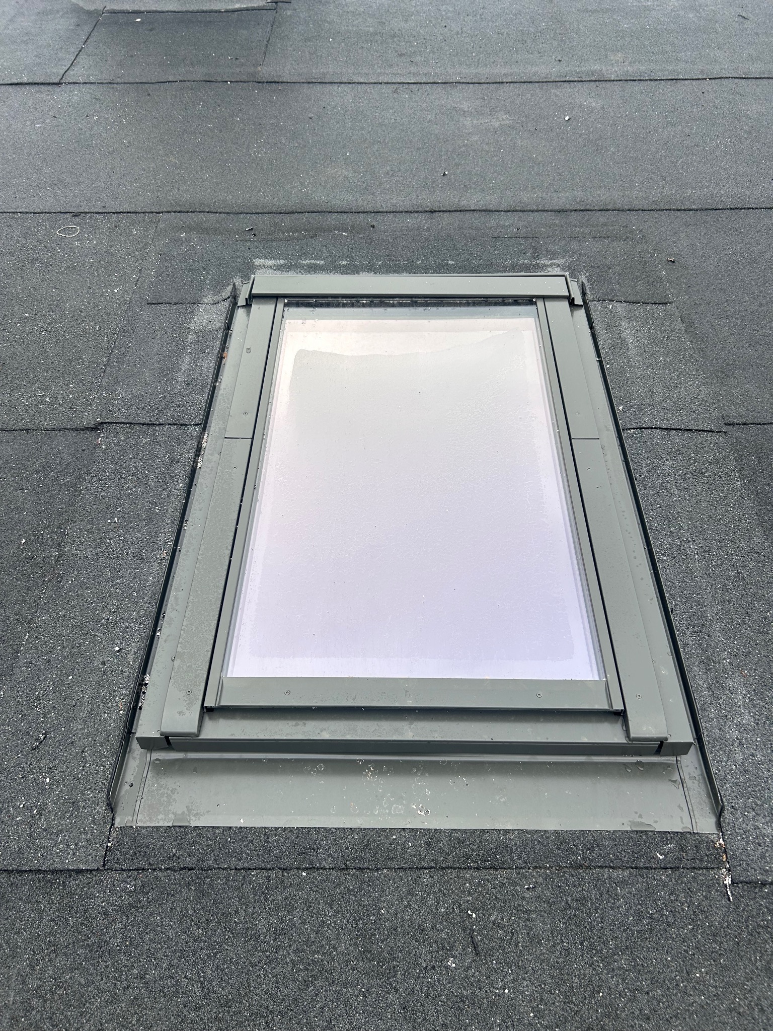 Szczelne okno dachowe z szarą ramą, wkomponowane w pokrycie z papy na płaskim dachu. Widoczne detale łączenia i uszczelnienia.