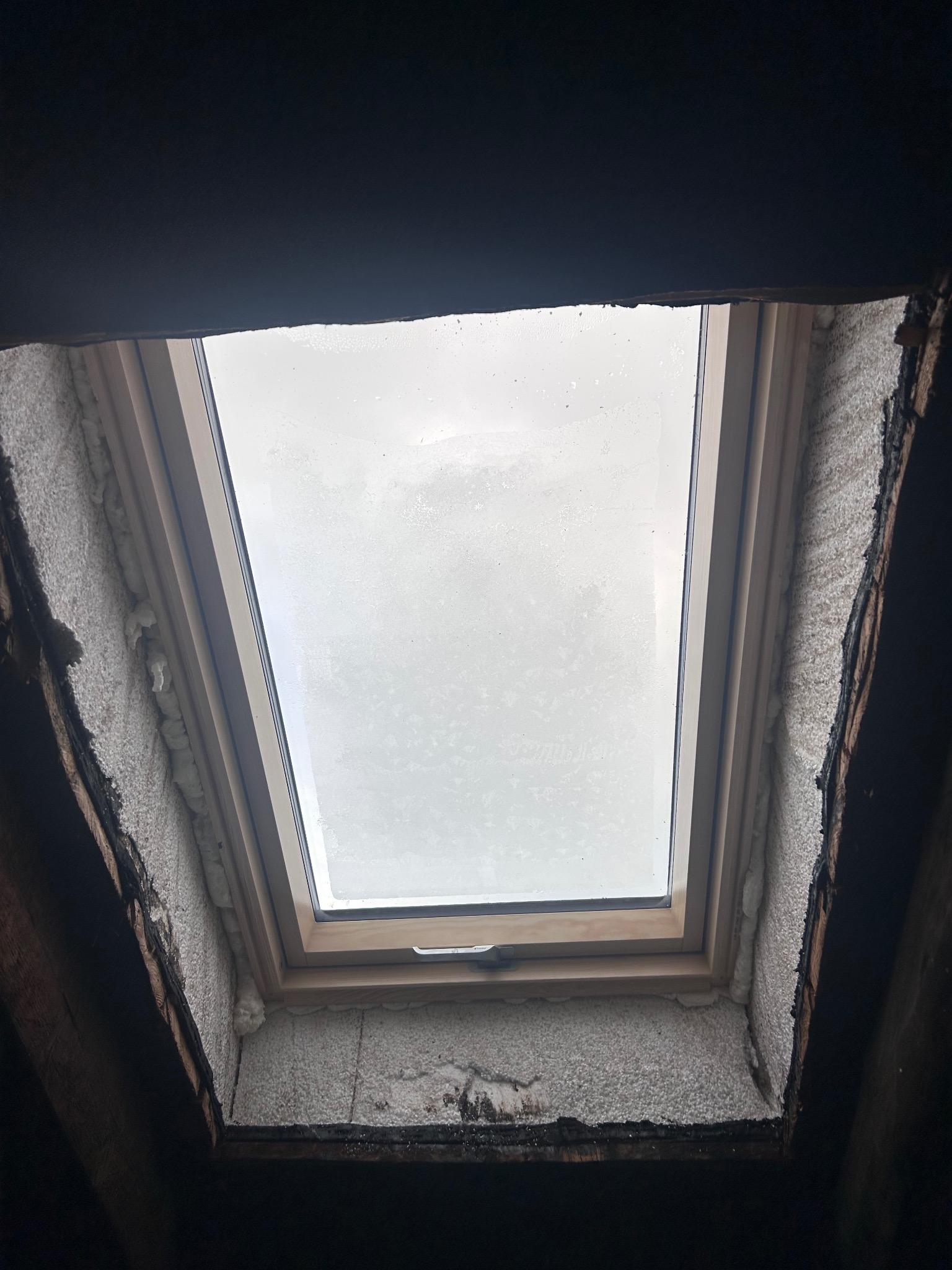 Zamontowane okno dachowe z widoczną pianką uszczelniającą, ujęcie od wewnątrz poddasza. Widoczne fragmenty konstrukcji dachu i niebo przez szybę.