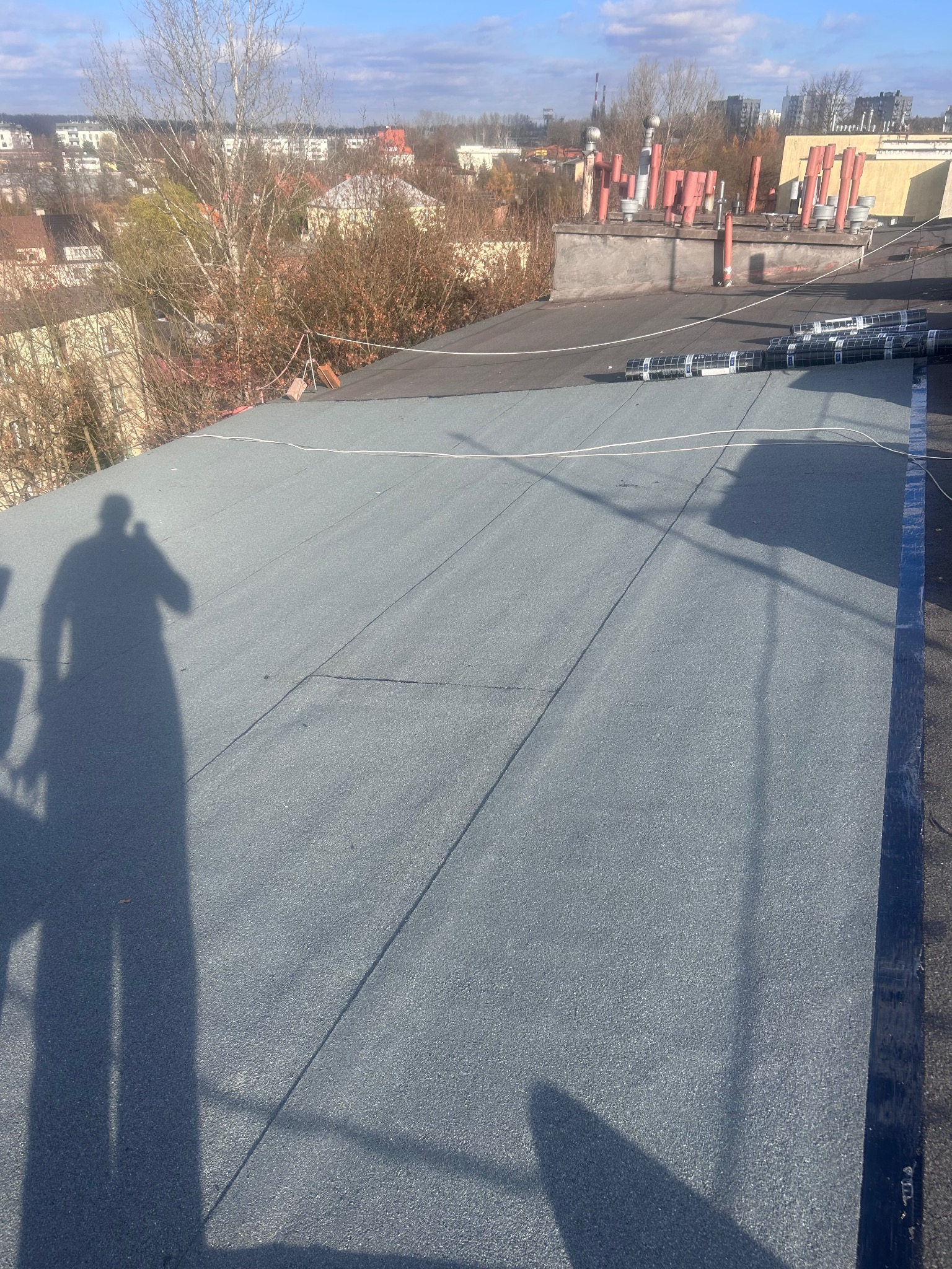 Szary, płaski dach pokryty papą termozgrzewalną w słoneczny dzień. W tle zabudowa miejska i cień osoby. Widoczne rolki papy przygotowane do montażu.