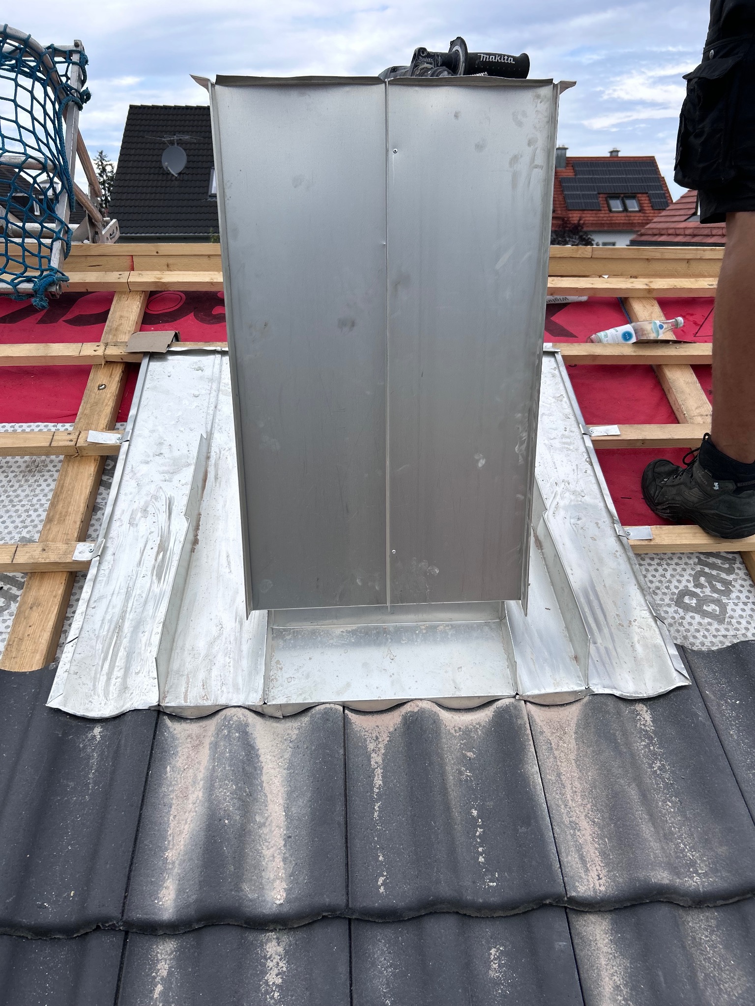 Montaż metalowego komina na dachu z czarnej dachówki. Widoczne drewniane elementy konstrukcji dachu i narzędzia. Ujęcie z bliska, podkreślające detale.