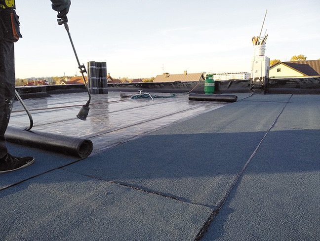 Praca na dachu: dekarz z palnikiem układa papę termozgrzewalną na płaskim dachu. Widoczne rolki papy i butla gazowa. Słoneczny dzień, widok na zabudowania w tle.