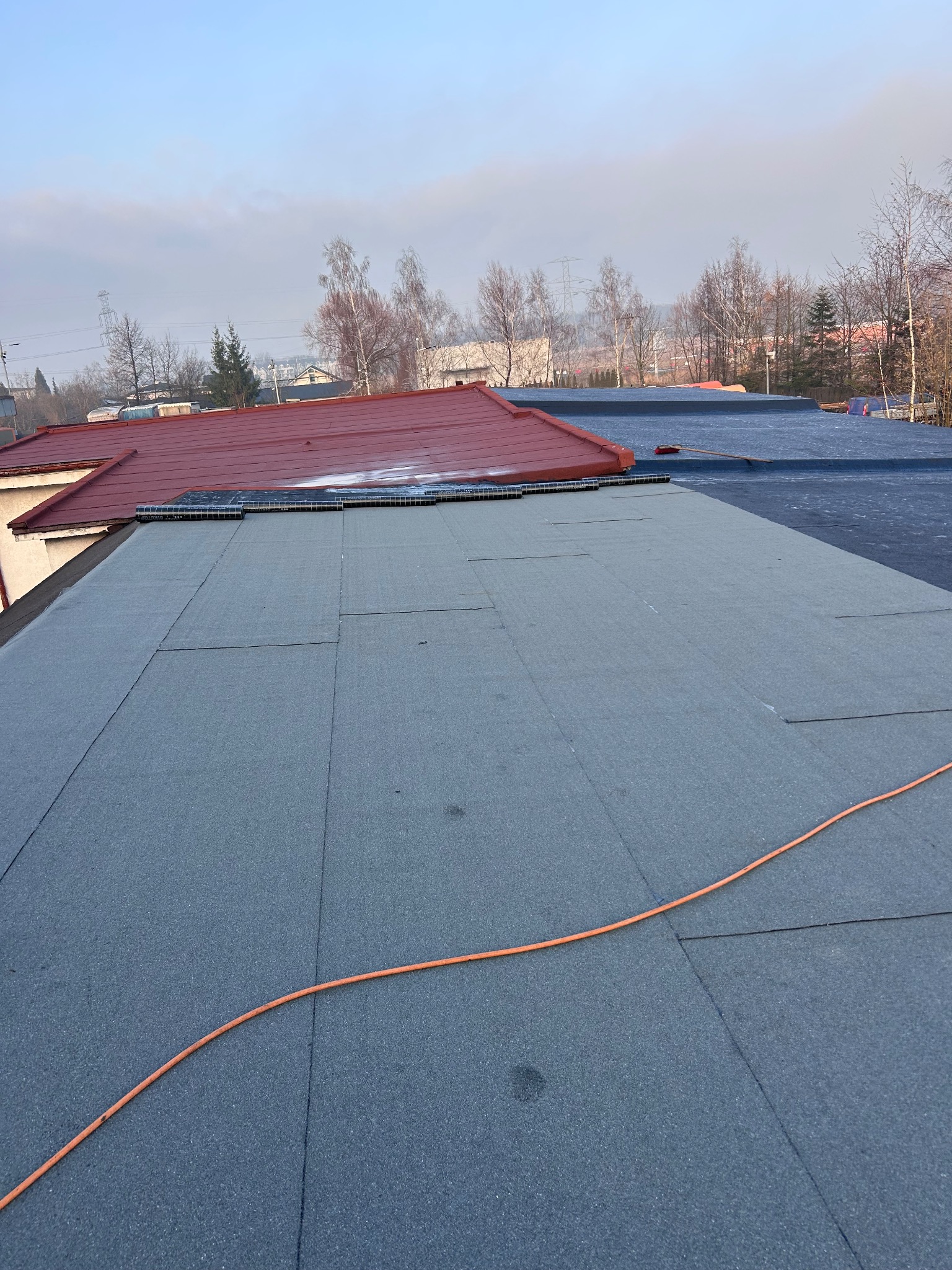 Fragment dachu pokrytego papą termozgrzewalną na płasko, z widocznym fragmentem dachu skośnego z czerwoną dachówką i pomarańczowym kablem na papie. W tle drzewa i słupy wysokiego napięcia.