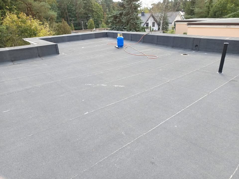 Szary, płaski dach pokryty papą termozgrzewalną z widocznym kominem wentylacyjnym, butlą gazową i zgrzewarką. W tle drzewa i budynki.