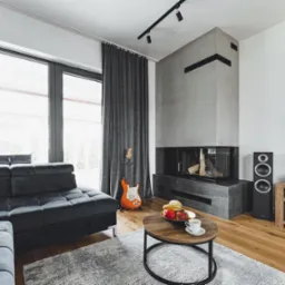 Nowoczesny salon z szarym narożnikiem, kominkiem obłożonym szarym kamieniem, gitarą elektryczną opartą o ścianę, okrągłym stolikiem kawowym z owocami i filiżanką, oraz dużym oknem z szarymi zasłonami.