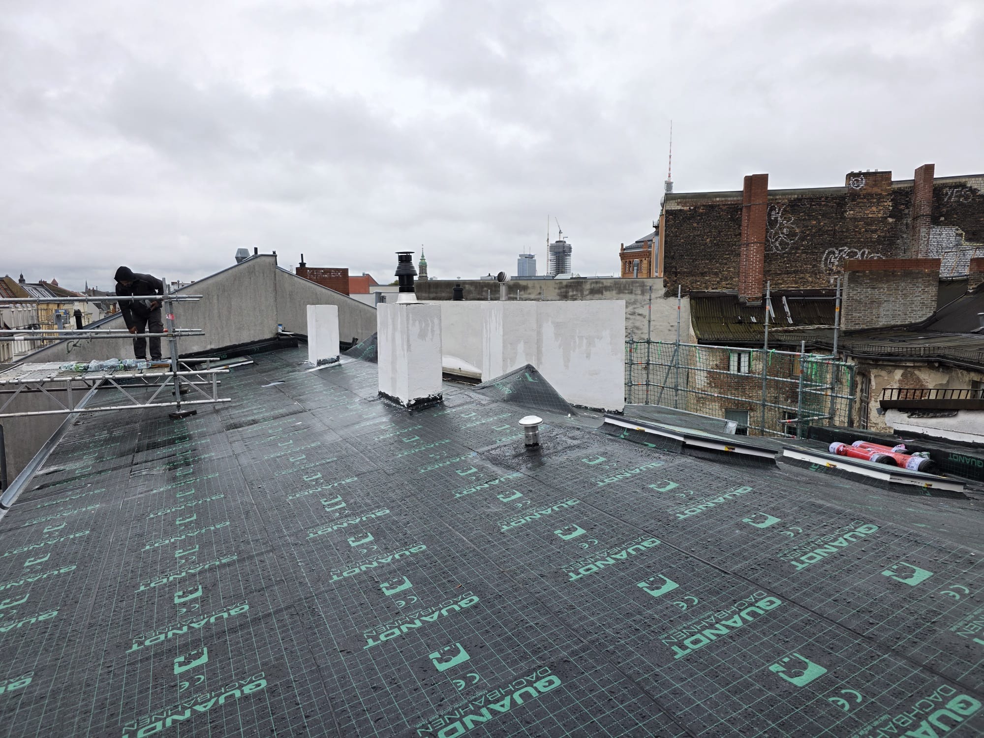 Układanie membrany dachowej Quantdt na dachu w Warszawie. Widoczny pracownik na rusztowaniu i elementy wentylacyjne. Prace dekarskie w toku, pochmurny dzień.
