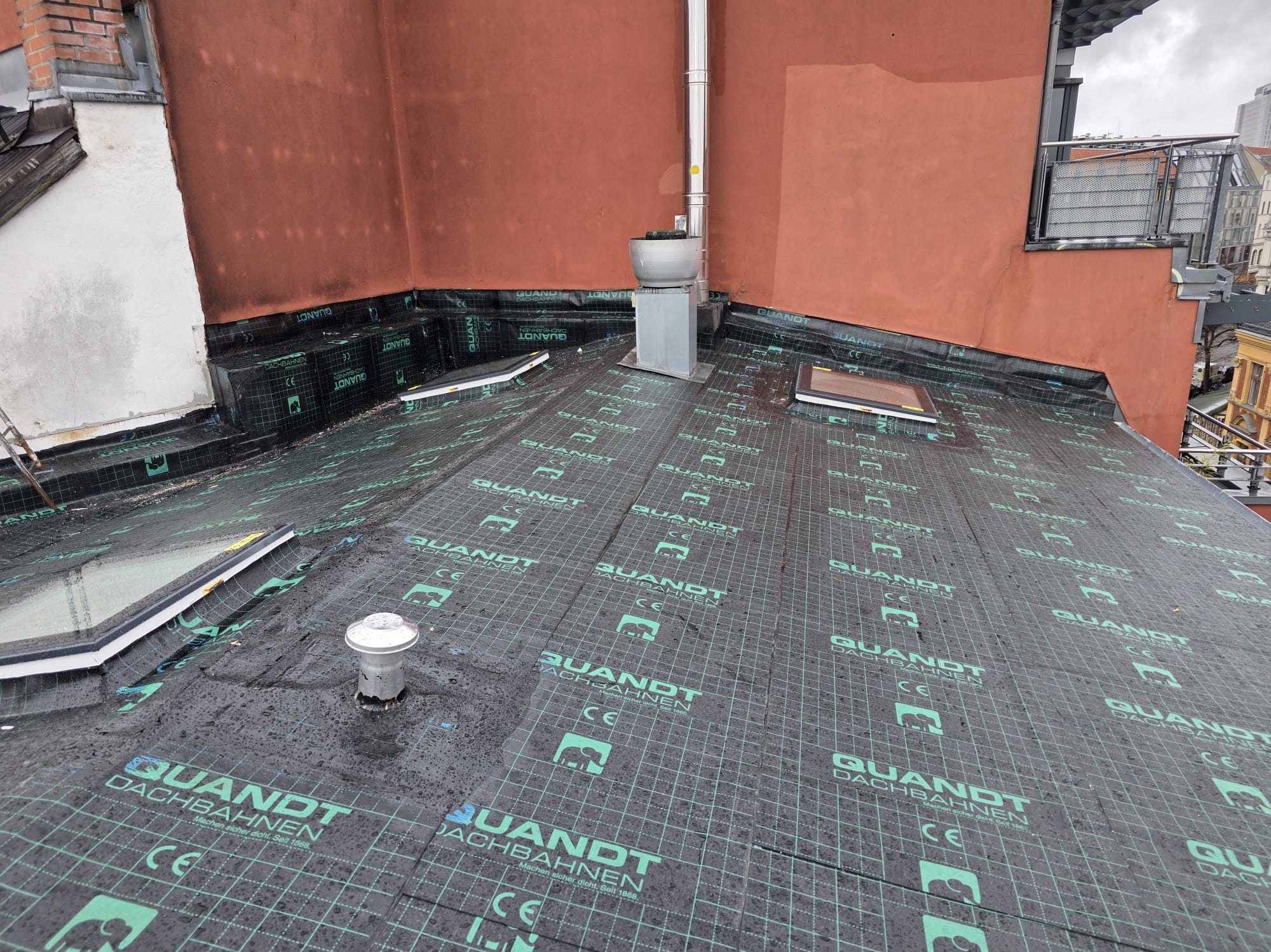 Dach pokryty membraną hydroizolacyjną z widocznymi detalami, takimi jak komin, okna dachowe i elementy wentylacyjne. Widok z góry na płaski dach z detalami architektonicznymi.