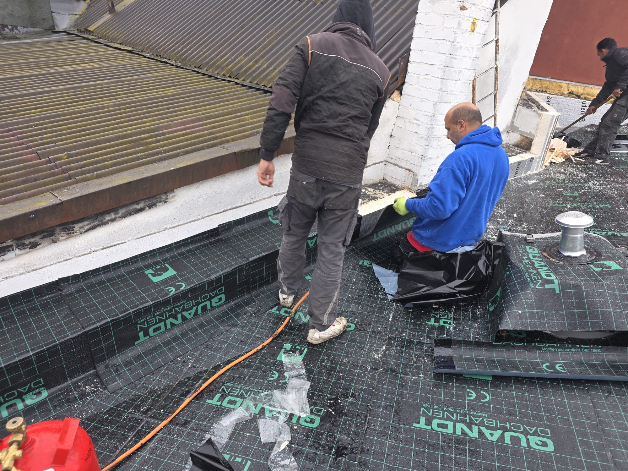 Montaż membrany dachowej QuantDT na dachu z blachy falistej. Widoczni dekarze podczas pracy, jeden siedzi, drugi stoi. Pomarańczowy kabel na membranie.