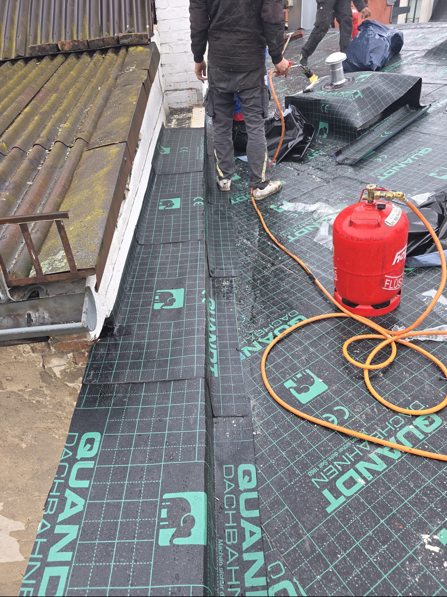 Prace dekarskie na dachu: ekipa montuje papę termozgrzewalną marki Quandt Dachbahnen, używając palnika i butli gazowej. Widoczny fragment starego dachu.