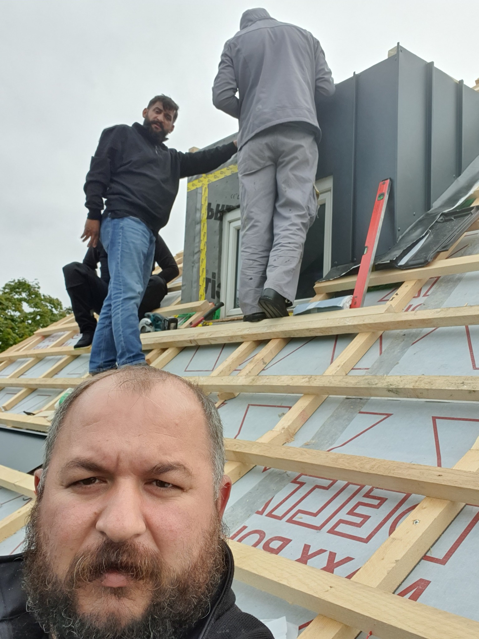 Ekipa dekarzy montuje obróbkę blacharską komina na dachu z drewnianą konstrukcją. Widoczny poziomica i narzędzia. Ujęcie z dołu, z perspektywy osoby na dachu.