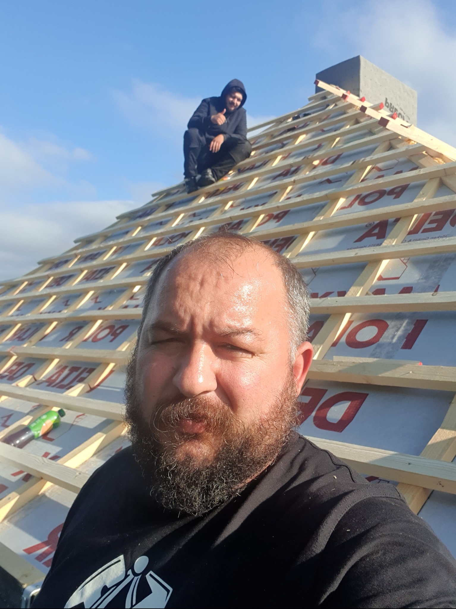 W trakcie budowy dachu: widoczna więźba dachowa z folią, na szczycie siedzi dekarz, a na pierwszym planie twarz drugiego dekarza.