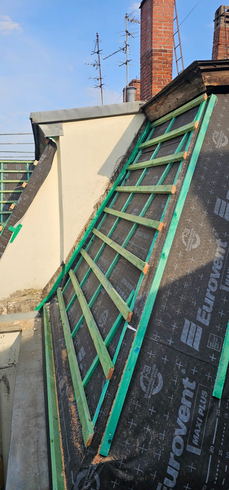 Dach z łatami i membraną Eurovent Maxi Plus, widok z góry, fragment komina w tle. Etap przygotowania do krycia dachu, jasne drewniane łaty na czarnej membranie.