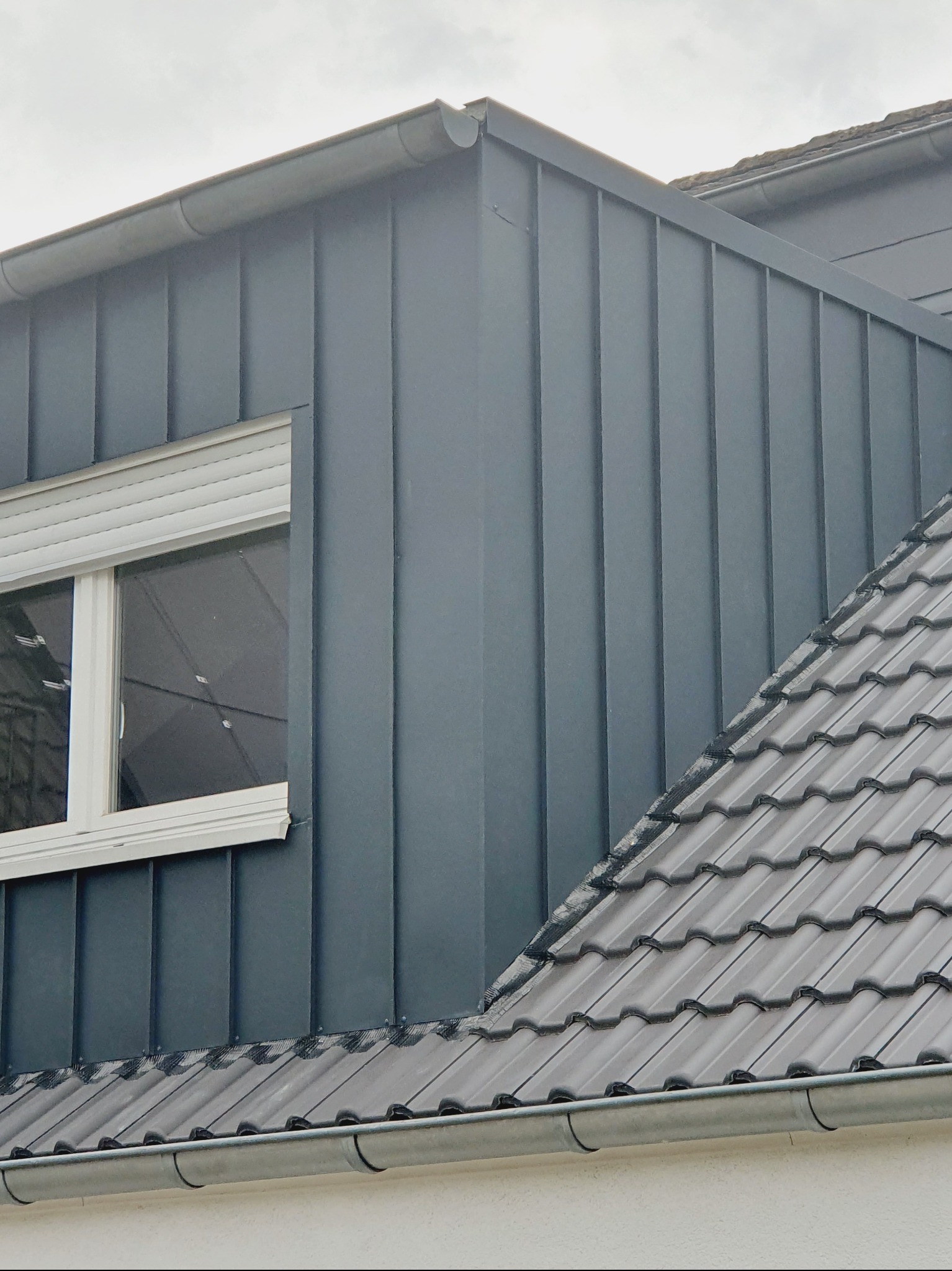 Szary dach z blachy na domu z oknem w Warszawie. Widoczna rynna i dachówka. Elewacja w pionowe pasy z blachy. Nowoczesne wykończenie.