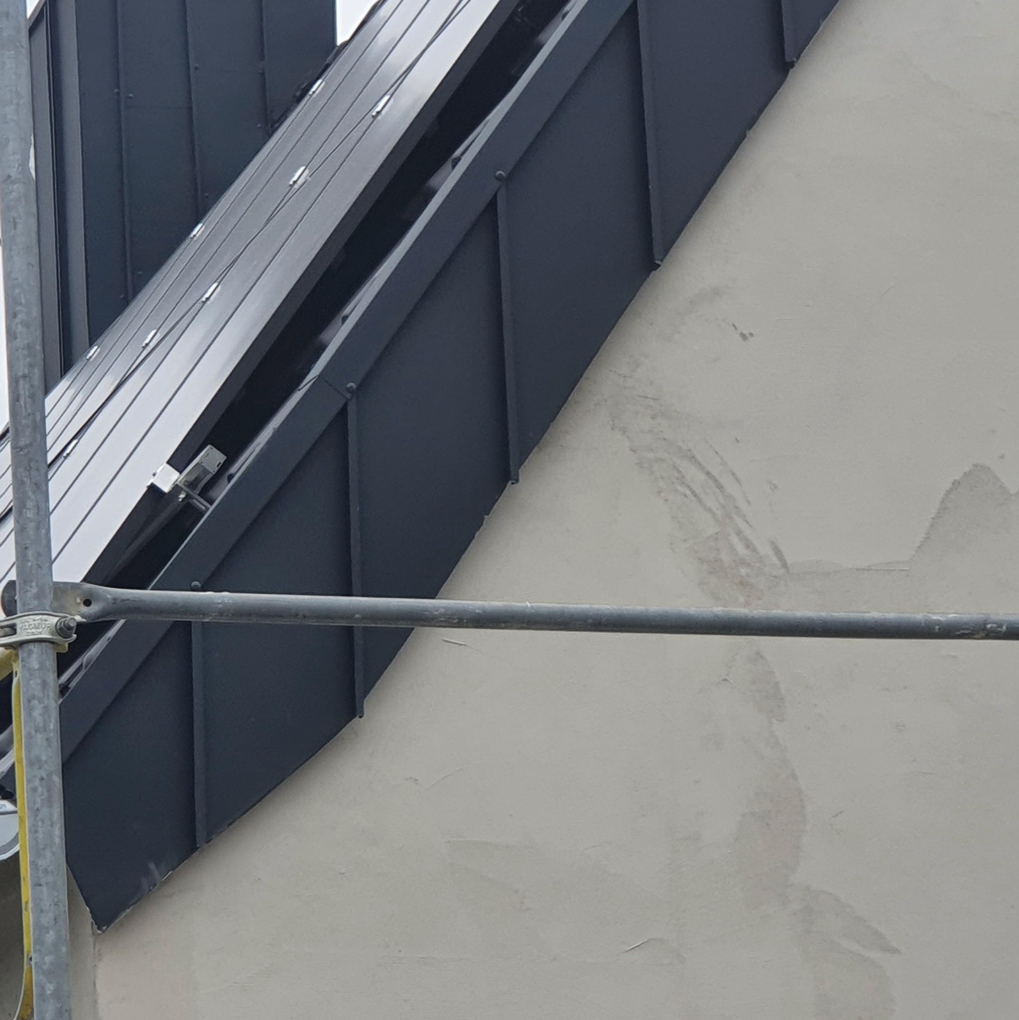 Fragment dachu z blachy na rąbek stojący w kolorze grafitowym, widoczny ze rusztowania. Ściana budynku w jasnym odcieniu. Detal łączenia dachu ze ścianą.