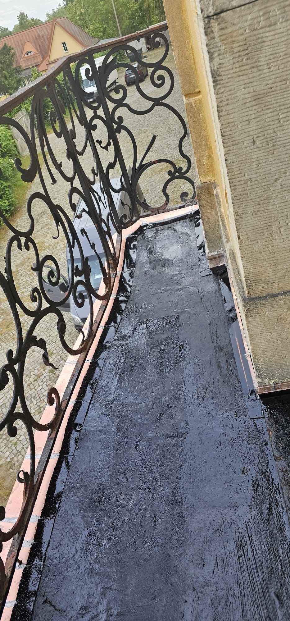 Świeżo zaizolowany, czarny dach balkonu z ozdobną, metalową balustradą i kamienną elewacją budynku w tle. Widok z góry, podkreślający detale izolacji.