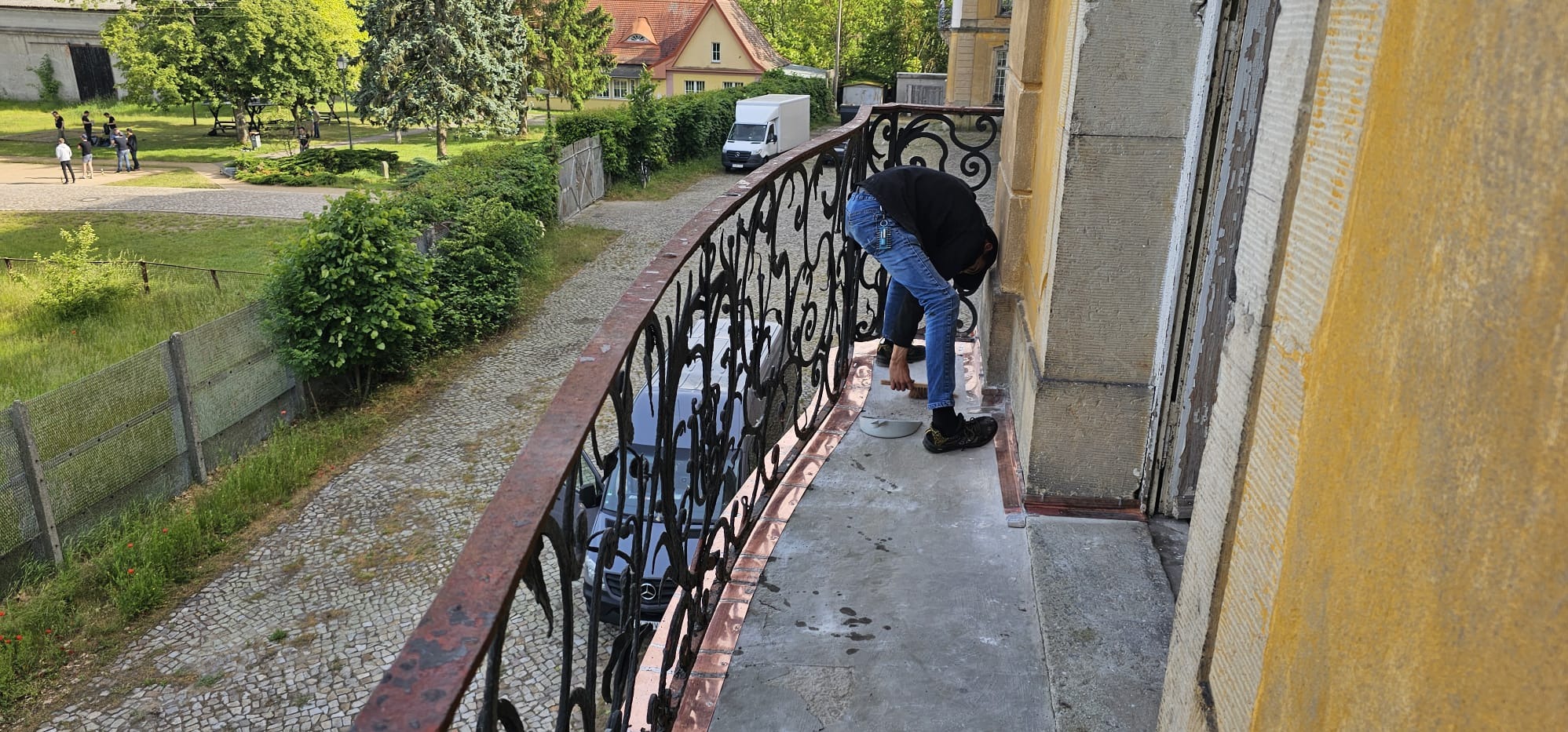 Pracownik czyści elewację budynku z balkonu, używając szczotki i wiadra. Widoczna ozdobna, metalowa balustrada balkonu i fragment elewacji w kolorze żółtym.
