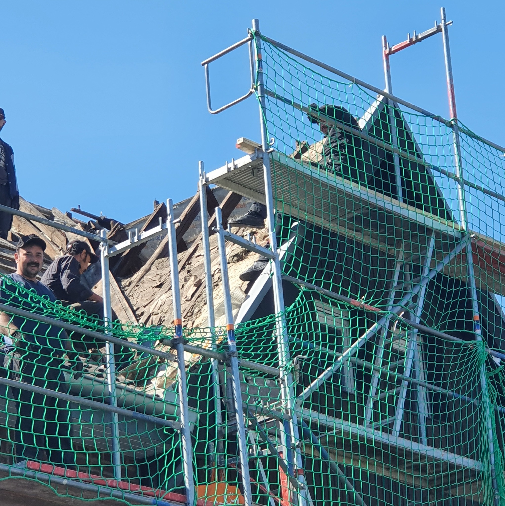 Ekipa dekarzy na rusztowaniu, zabezpieczona siatką, pracuje nad wymianą poszycia dachu. Widoczne stare deski i fragment nieba. Jeden z pracowników uśmiecha się.