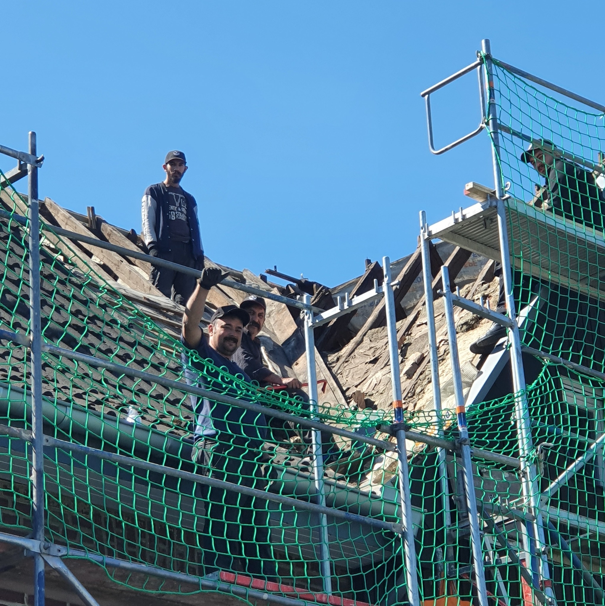 Ekipa dekarzy na rusztowaniu, z zadowoleniem prezentuje postęp prac przy wymianie starego dachu. Widoczne uszkodzone elementy konstrukcji i błękitne niebo w tle.
