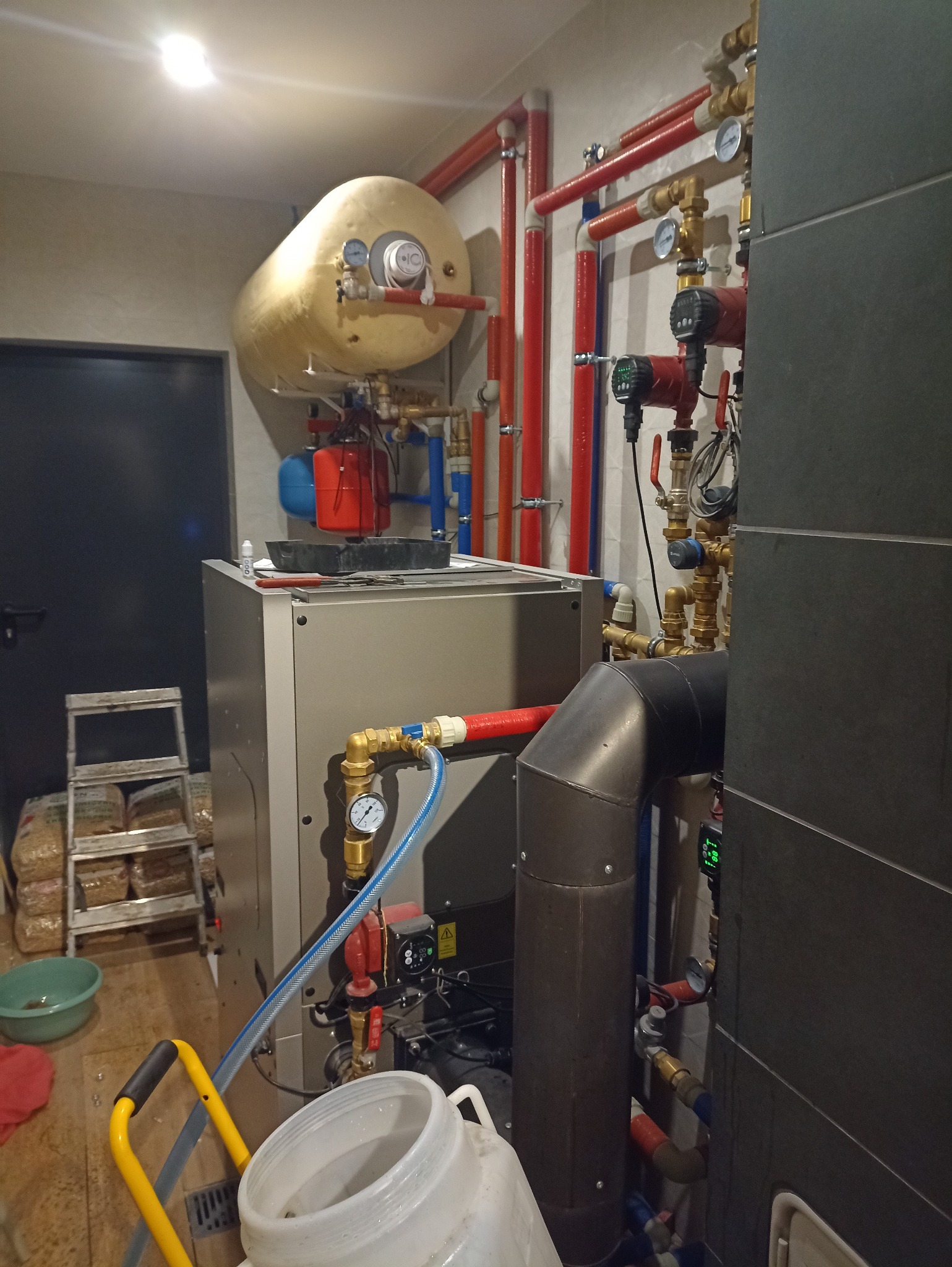 Kotłownia z piecem na pellet, zasobnikiem ciepłej wody i instalacją grzewczą z rurami miedzianymi i plastikowymi. Widoczne manometry i pompy obiegowe.