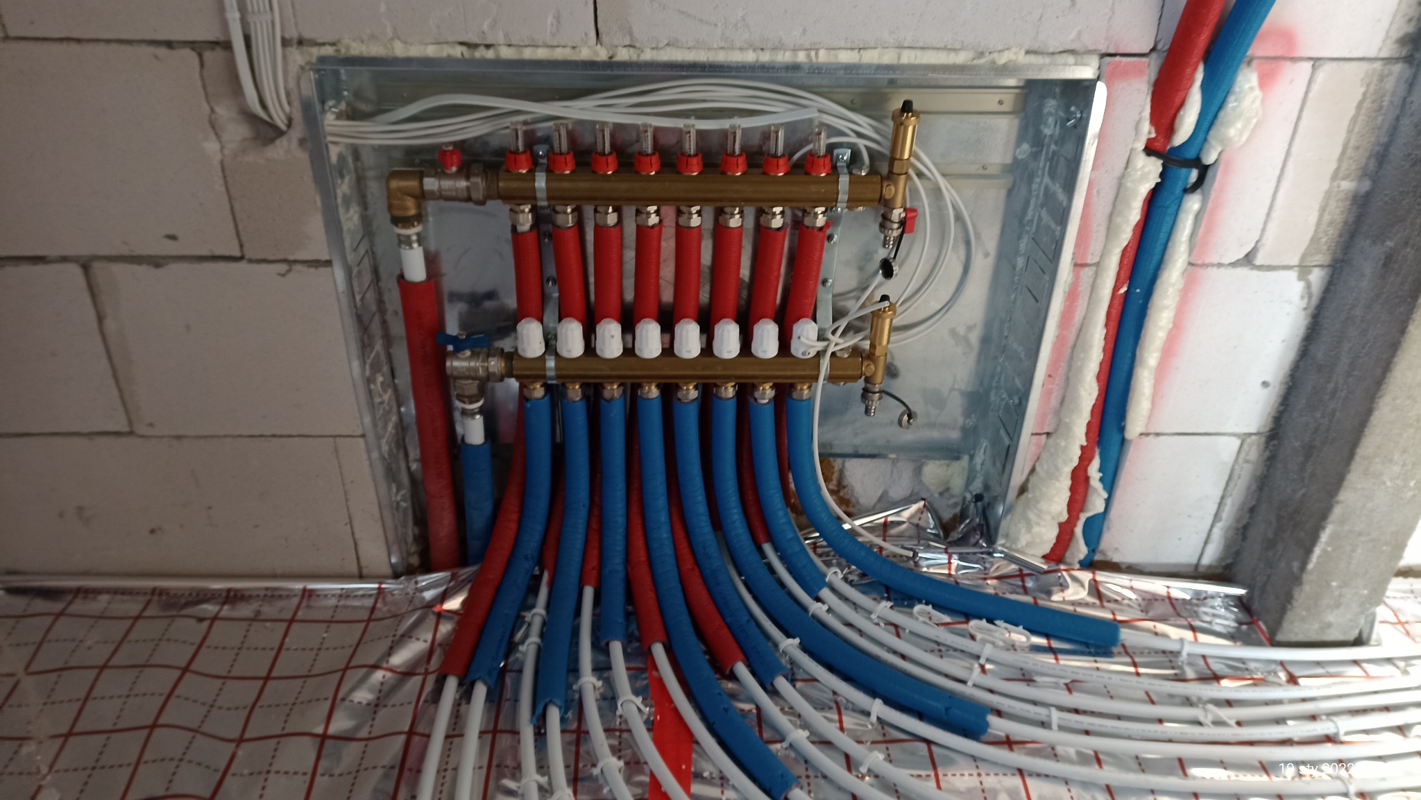 Rozdzielacz ogrzewania podłogowego z rurami w izolacji czerwonej i niebieskiej, zamontowany na ścianie z bloczków, widoczne białe przewody i folia izolacyjna na podłodze.