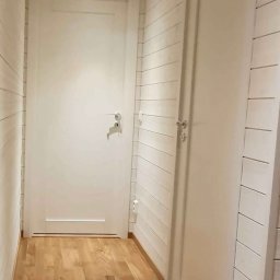 Jan Trąbka - Jasne wnętrze z drewnianą podłogą i boazerią. Dwoje białych drzwi, jedne z klamką i zamkiem, drugie gładkie. Minimalistyczny design, ciepłe oświetlenie.