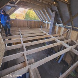 Jan Trąbka - Drewniana konstrukcja podłogi na poddaszu w trakcie budowy, widok z góry. Fragment komina i pracownik w tle. Naturalne światło.