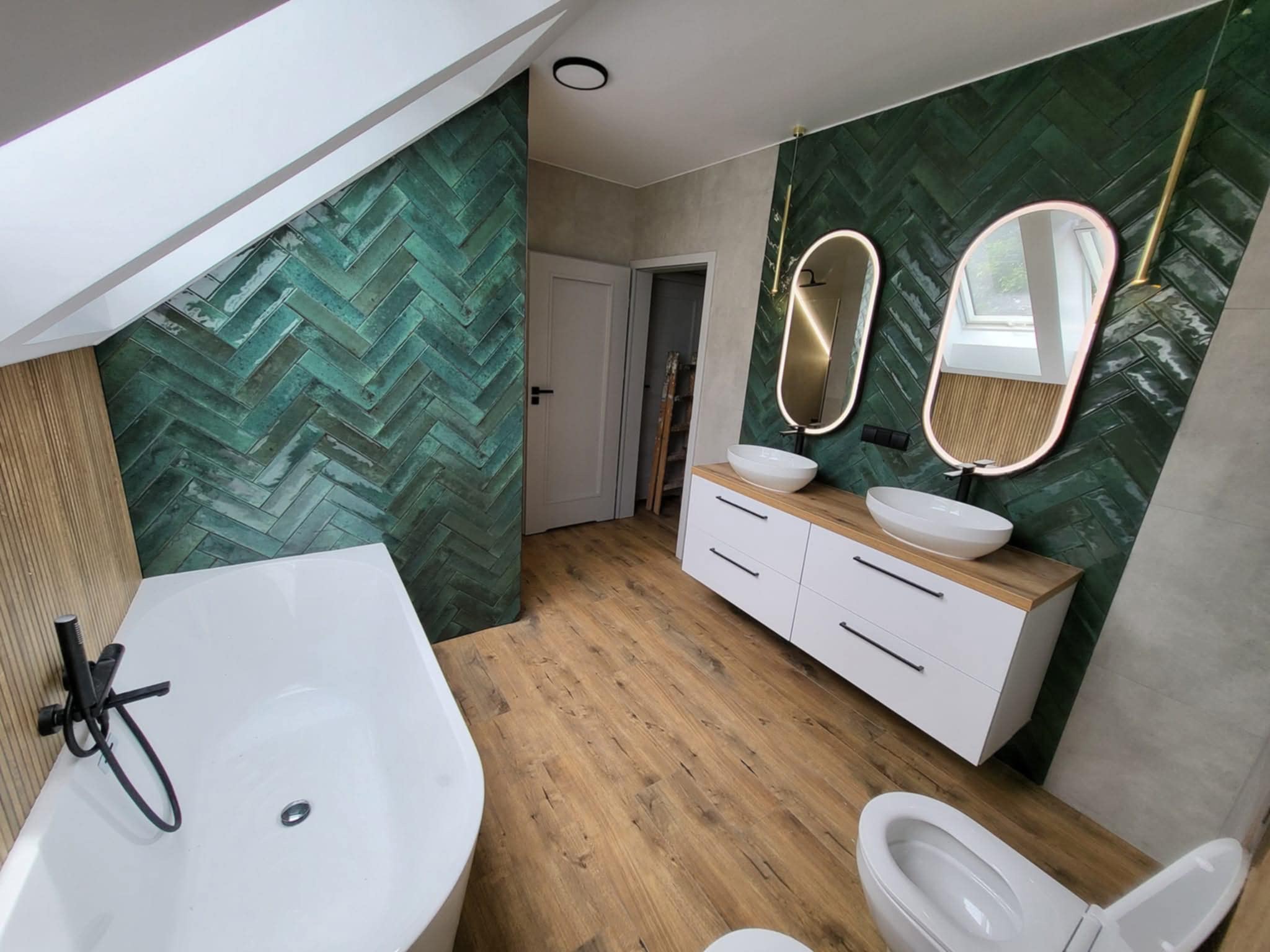 Nowoczesna łazienka na poddaszu z zieloną ścianą w jodełkę, drewnianą podłogą i białymi meblami. Uwagę przykuwają owalne lustra z podświetleniem LED.