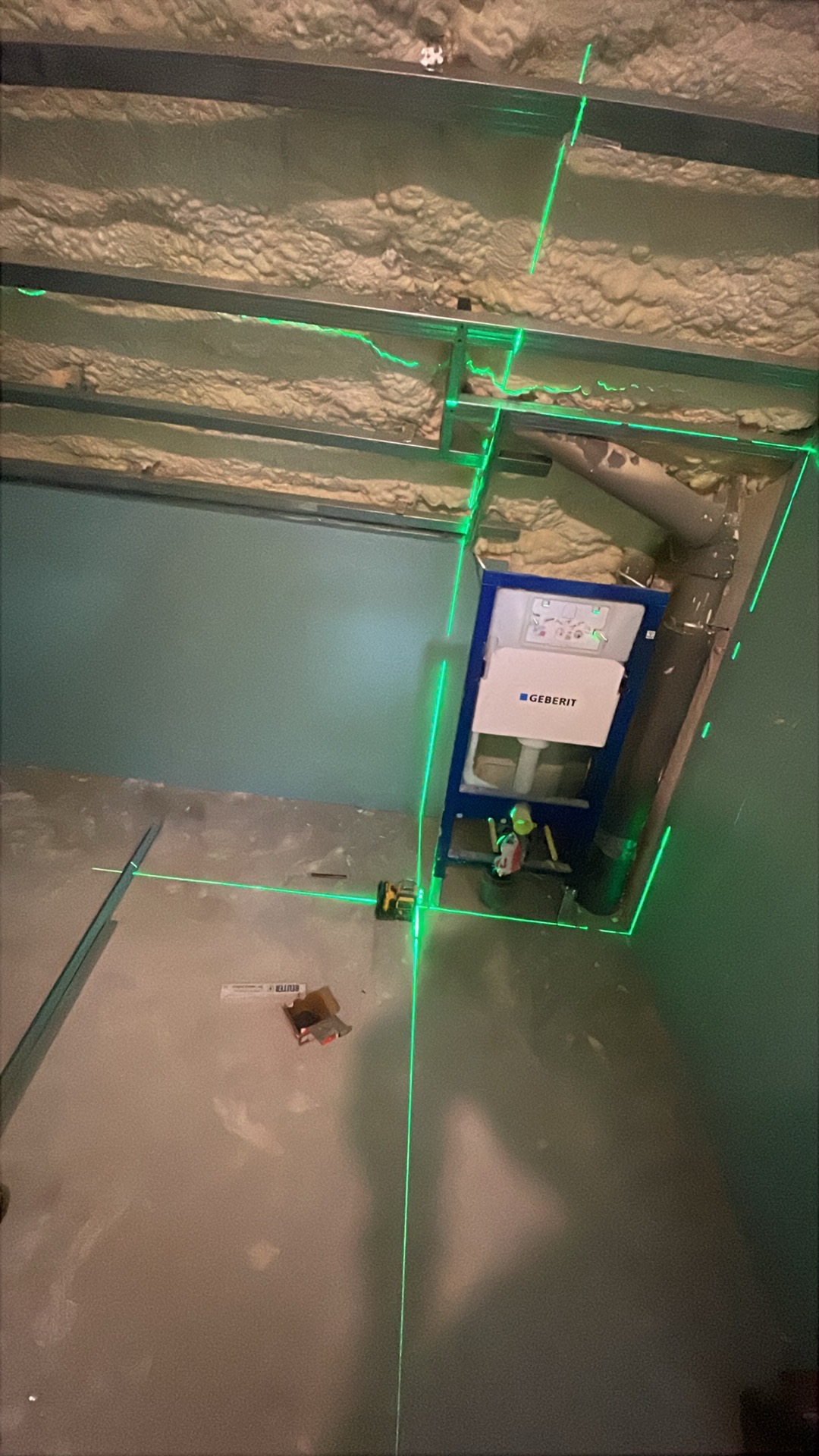 Montaż stelaża WC Geberit na tle surowego wnętrza łazienki z widoczną izolacją i metalową konstrukcją sufitu podwieszanego, wyznaczony poziom lasera.