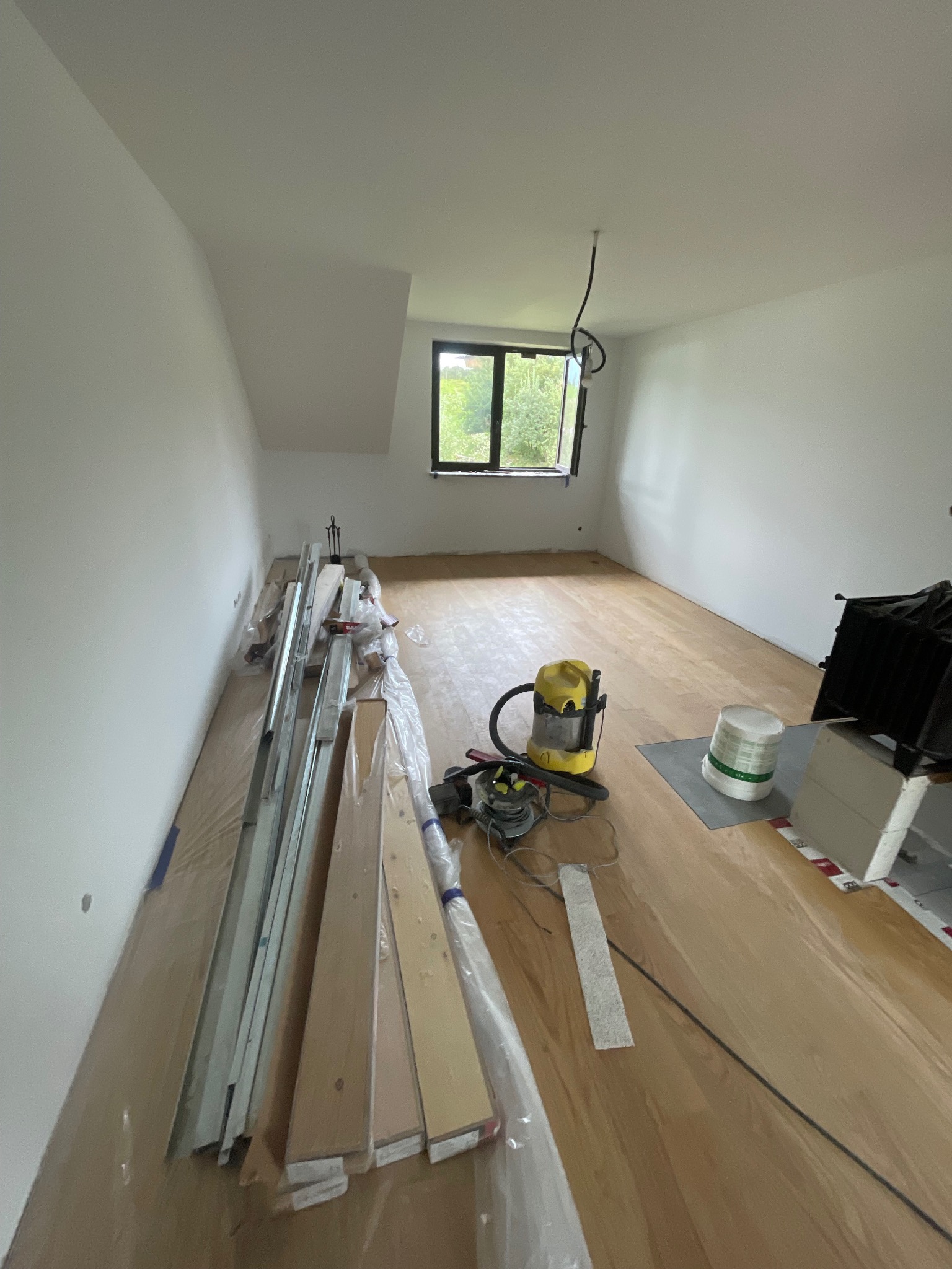 Jasne wnętrze w trakcie układania drewnianej podłogi. Widoczne panele, listwy przypodłogowe, narzędzia i odkurzacz. Okno z widokiem na zieleń.