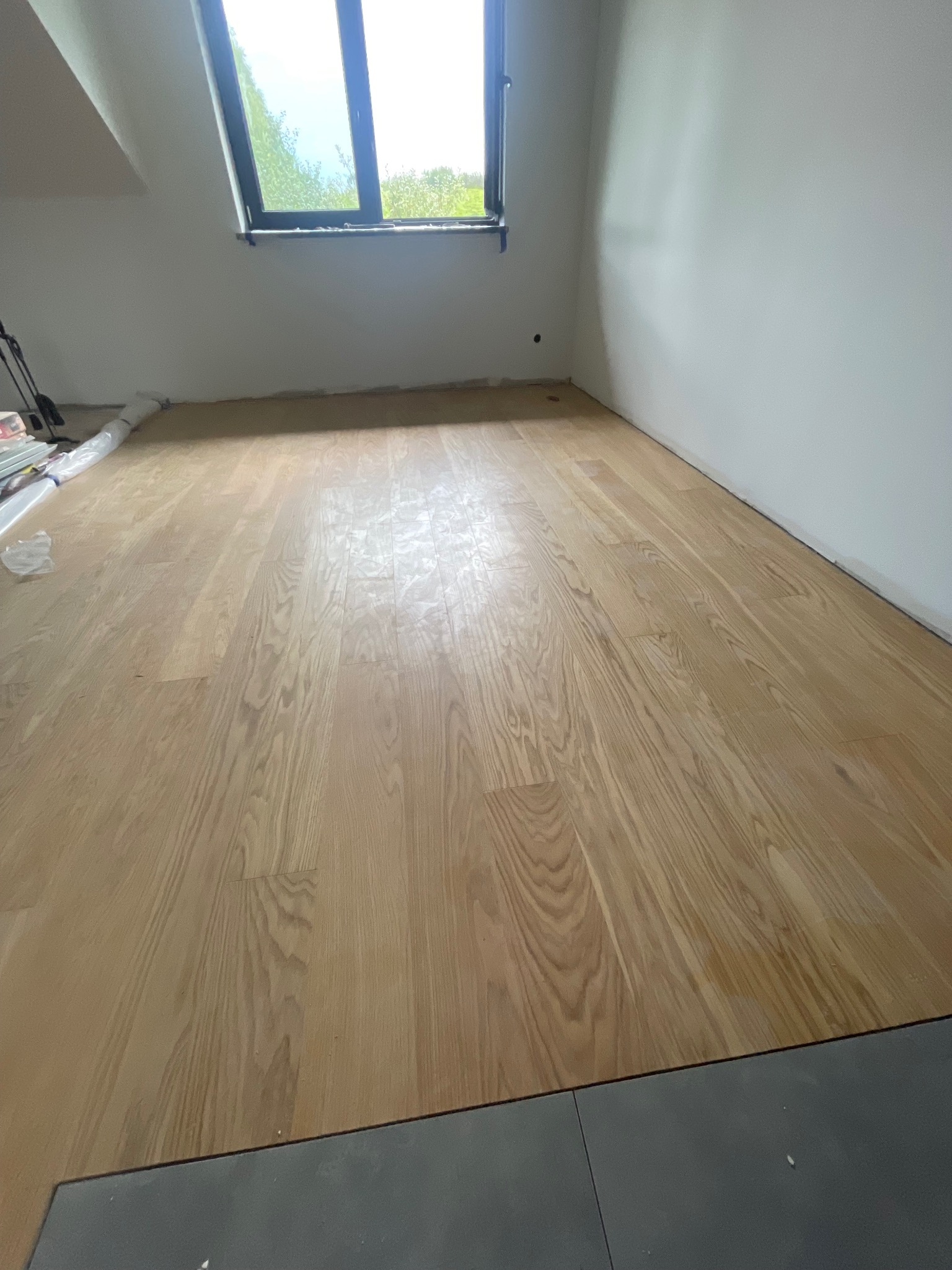 Jasne panele podłogowe ułożone w pokoju z oknem dachowym, widoczne materiały budowlane w rogu. Szara płytka na styku z panelami.