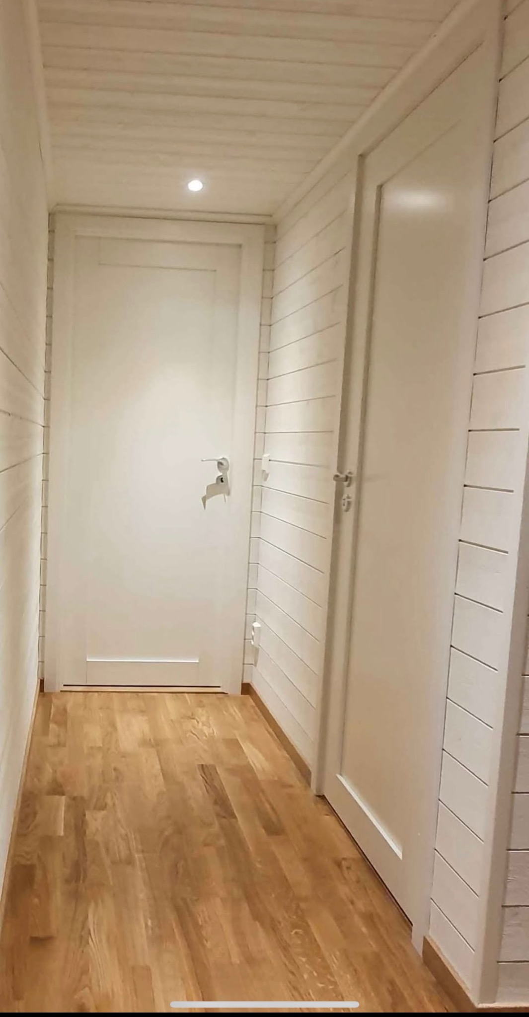 Jasne wnętrze z drewnianą podłogą i boazerią. Dwoje białych drzwi, jedne z klamką i zamkiem, drugie gładkie. Minimalistyczny design, ciepłe oświetlenie.