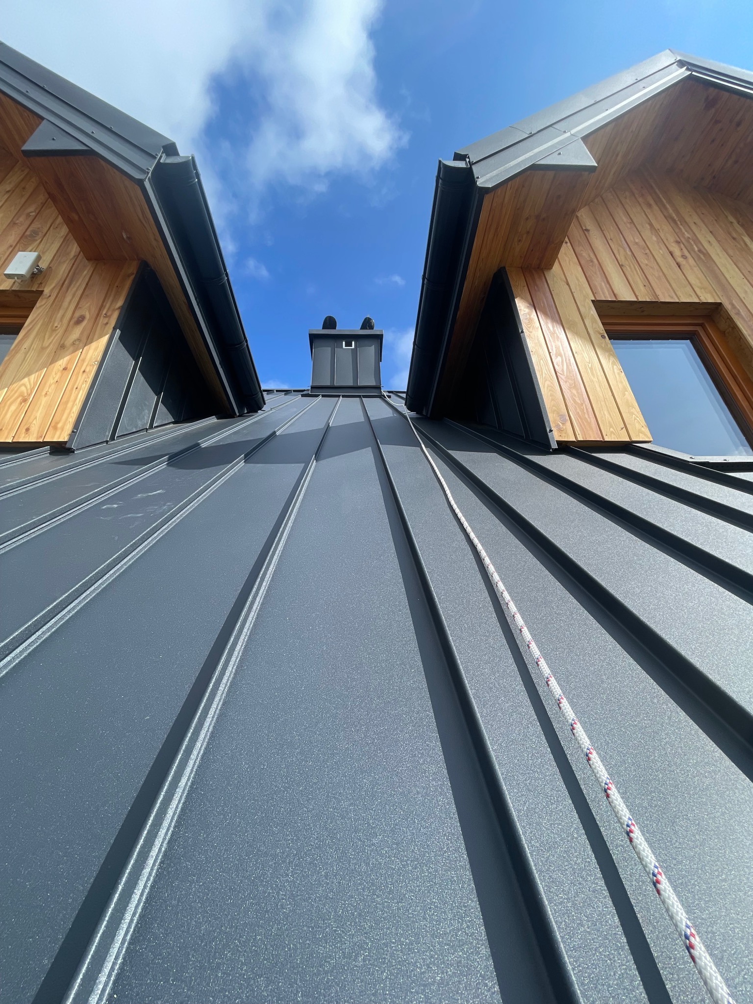 Szary dach z blachy trapezowej, widok z dołu na komin i fragmenty drewnianej elewacji pod błękitnym niebem. Widoczna lina zabezpieczająca.