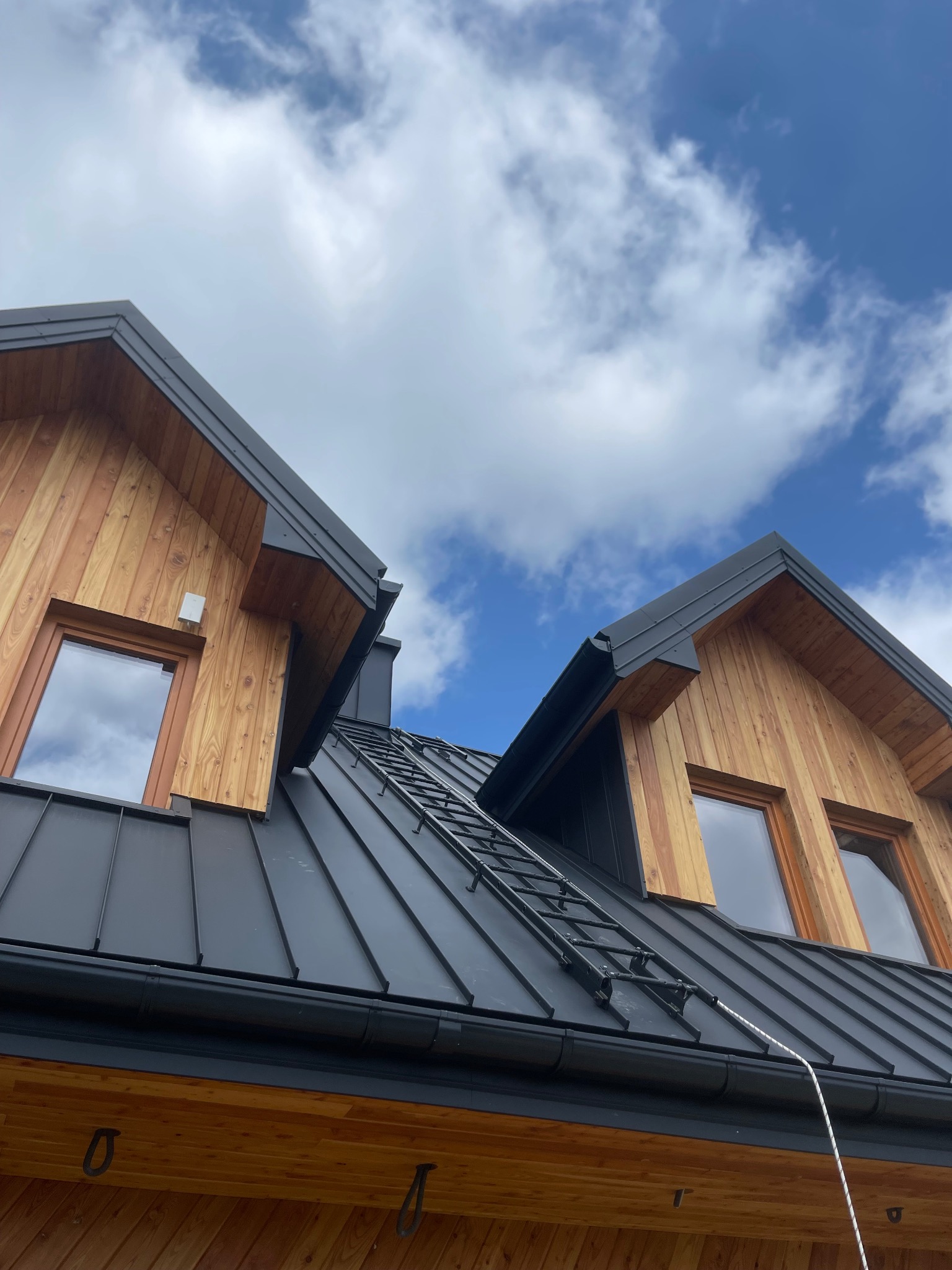 Nowoczesny dom z drewnianą elewacją i czarnym dachem z drabinką dachową, widok z dołu na tle błękitnego nieba z chmurami.