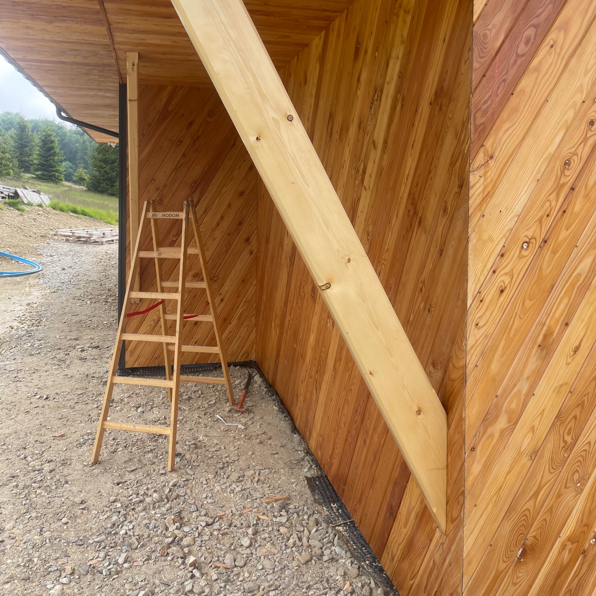 Ściana z desek ułożonych pod kątem, drewniana drabina oparta o ścianę, widoczne elementy konstrukcyjne. Prace wykończeniowe w drewnie.