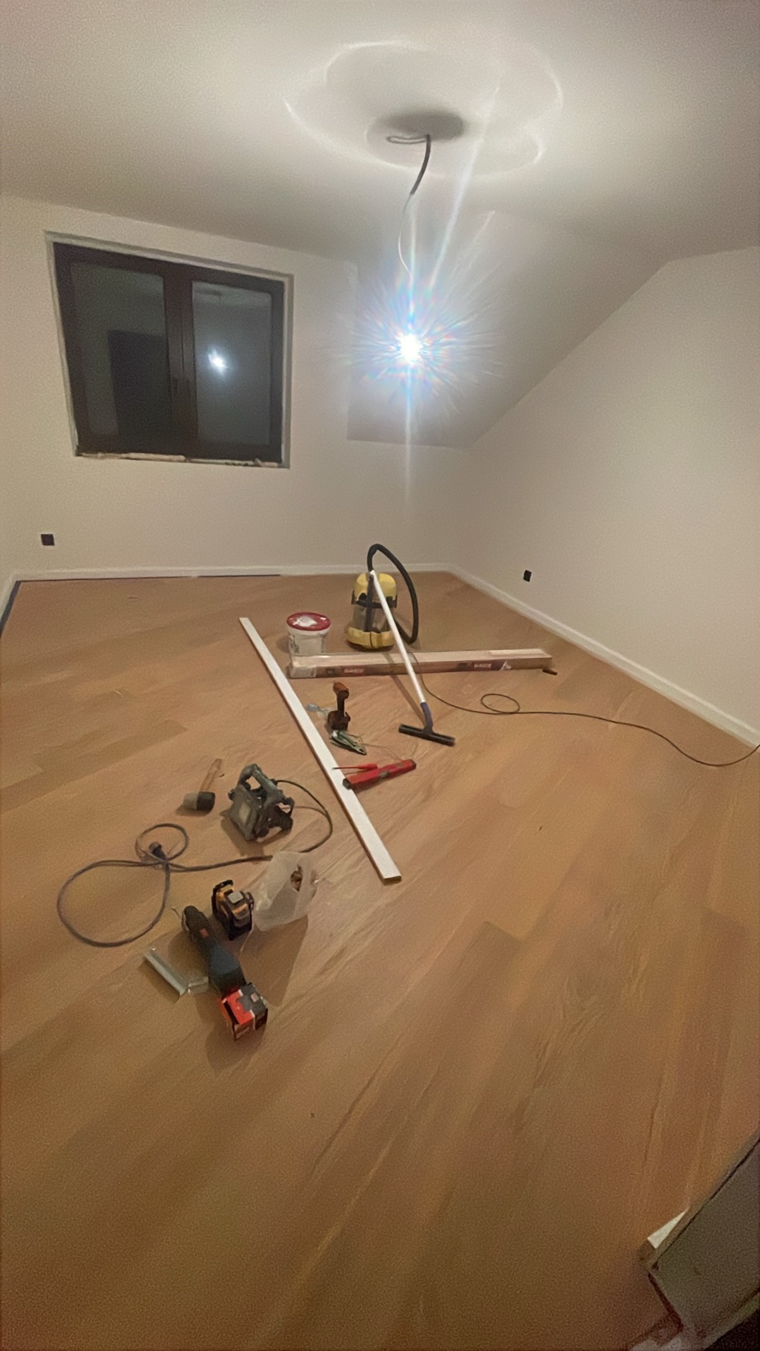 Jasne panele podłogowe w pokoju na poddaszu, przygotowane do montażu listwy przypodłogowej. Widoczne narzędzia i odkurzacz. Okno w tle.