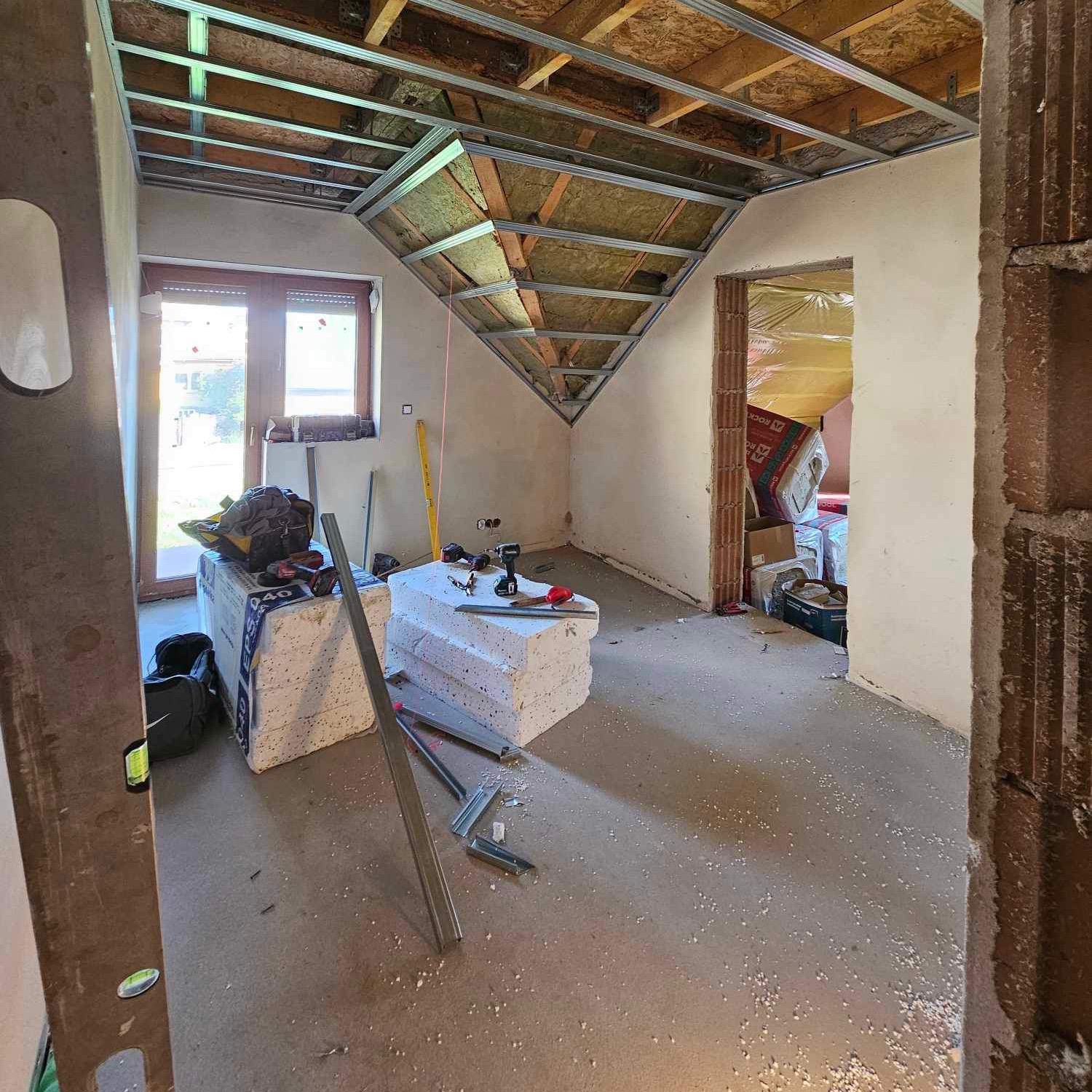 Skośne poddasze w trakcie adaptacji: metalowa konstrukcja sufitu podwieszanego, izolacja, narzędzia, poziomica. Widok na okno i przygotowane wejście do pomieszczenia.