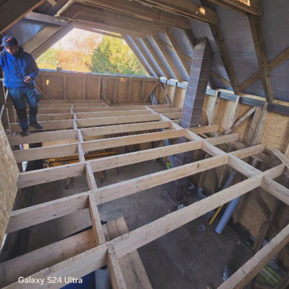 Drewniana konstrukcja podłogi na poddaszu w trakcie budowy, widok z góry. Fragment komina i pracownik w tle. Naturalne światło.