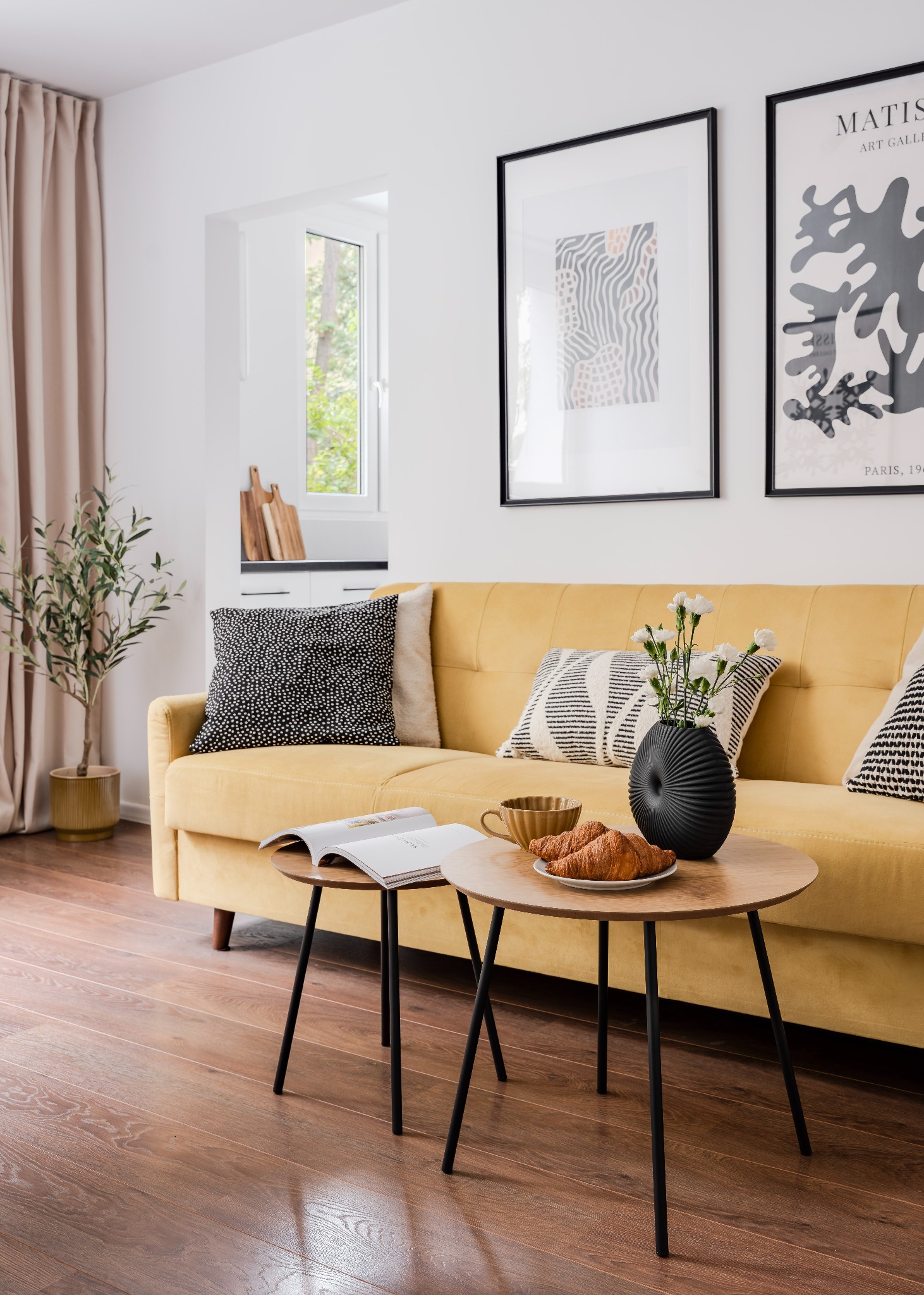 Żółta sofa z poduszkami w salonie. Na stoliku kawa, rogaliki i wazon z kwiatami. Na ścianie grafiki w czarnych ramkach. Jasne wnętrze z drewnianą podłogą.
