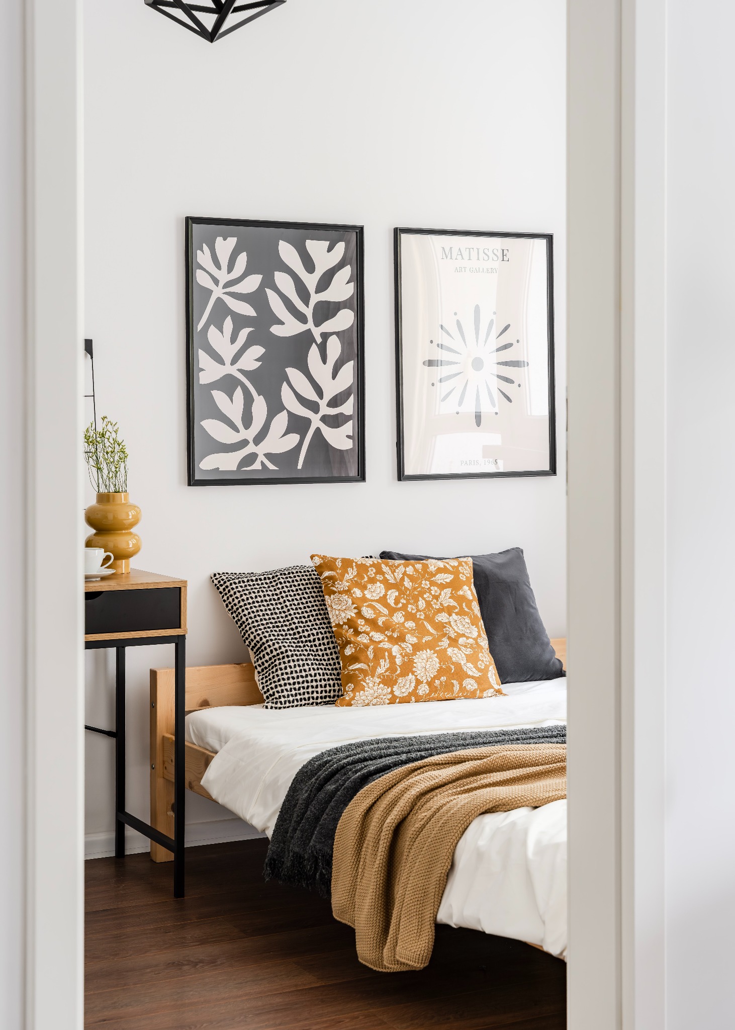 Jasna sypialnia z drewnianym łóżkiem, wzorzystymi poduszkami i plakatami w czarnych ramach. Na stoliku nocnym wazon z kwiatami i filiżanka.