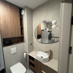 Przemysław Kieliński Remonty i Wykończenia - Nowoczesna łazienka z wiszącą toaletą, umywalką nablatową i szafką z drewnianym fornirem. W lustrze odbija się pralka i suszarka.