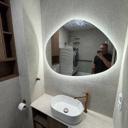 Przemysław Kieliński Remonty i Wykończenia - Nowoczesna łazienka z umywalką nablatową, baterią w kolorze miedzi i podświetlanym lustrem o nieregularnym kształcie. W tle widoczna pralka i kabina prysznicowa.