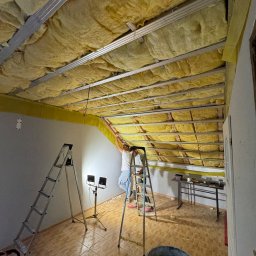 Przemysław Kieliński Remonty i Wykończenia - Montaż izolacji stropu wełną mineralną, widoczne profile metalowe, drabiny i narzędzia. Prace wykończeniowe w pomieszczeniu z terakotą na podłodze.