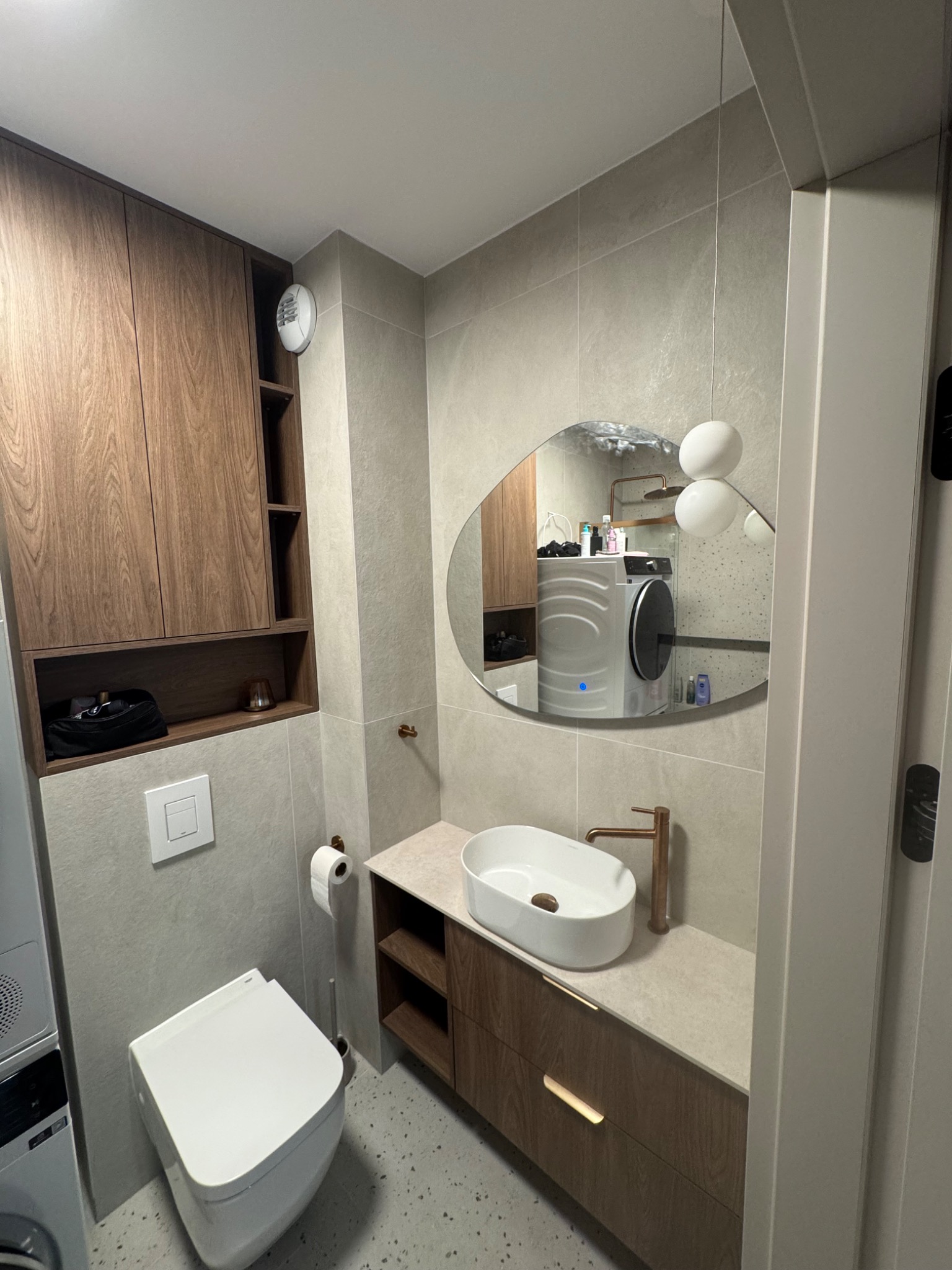 Nowoczesna łazienka z wiszącą toaletą, umywalką nablatową i szafką z drewnianym fornirem. W lustrze odbija się pralka i suszarka.