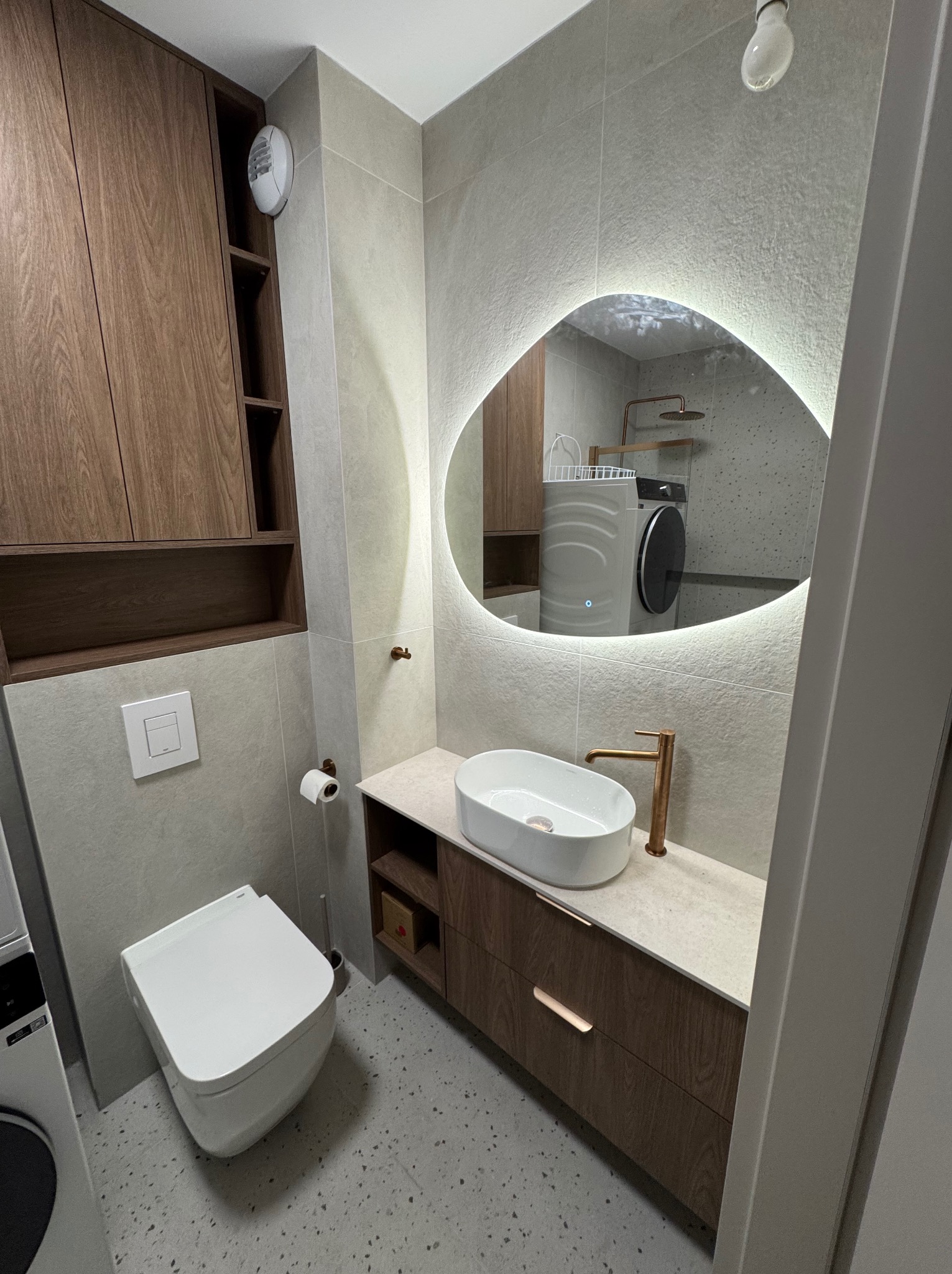 Nowoczesna łazienka z umywalką nablatową, podświetlanym lustrem o nieregularnym kształcie i drewnianymi szafkami. W tle widoczna pralka i prysznic.